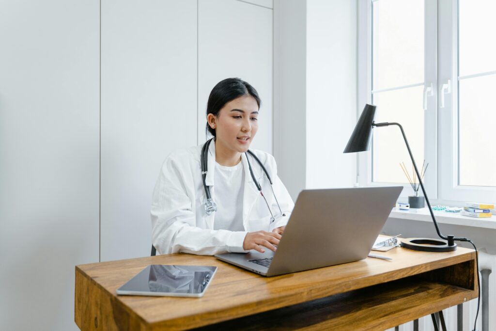 pexels-photo-8376232-8376232 A female doctor in a white coat uses a laptop for an online consultation from her office.